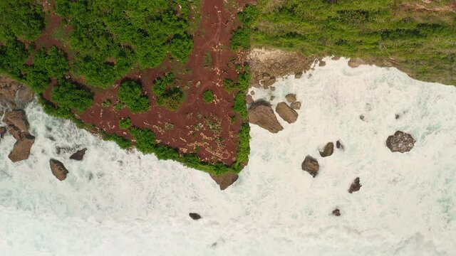 Aerial view of ocean blue waves break on high cliff of a rocky mountain. Danger sea waves on a beach aerial view drone shot. Bird's eye view of ocean waves crashing on a big stones on empty stone.
