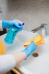 Close up of woman hands in protective gloves cleaning bathroom faucet tap and basin with cloth and sanitizer spray. Hygiene concept.