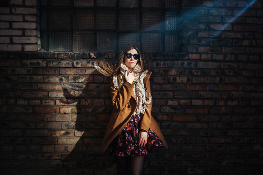 a young beautiful girl in sunglasses and a trench coat stands in a beam of light against a brick wall