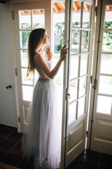 Fototapeta na wymiar a young beautiful girl in a white dress who looks like a bride is walking looks through the open glass door and is happy in the morning