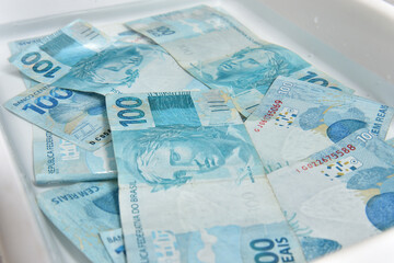 Notas de cem reais sendo lavadas com &aacute;gua numa forma de pl&aacute;stico branca