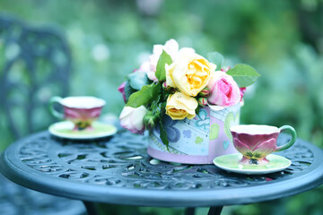 A bouquet of garden roses and two porcelain cups on a vintage table in the garden. A nice sitting area in the garden. soft selective gentle focus.