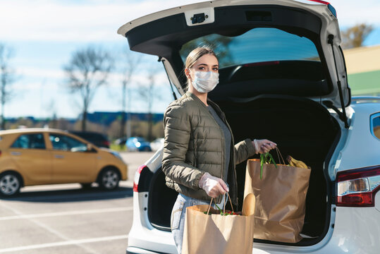 A young woman taking groceries from a supermarket from the car trunk. Social distancing: face mask, disposable gloves to prevent infection. Food shopping during coronavirus Covid-19 quarantine