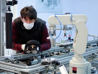 Man with surgical mask on face against COVID-19 ( Coronavirusis ) programming robot arm with control panel which is integrated on smart factory production line. industry 4.0 automation line.
