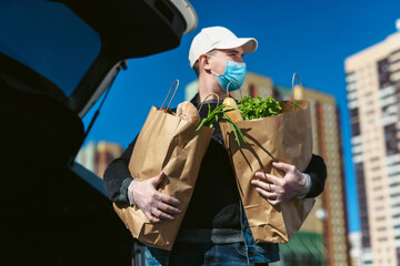 A courier in a protective mask and medical gloves delivers grocery bags from the supermarket. Auto trunk in the background. Social distancing. Coronavirus Covid 19 quarantine delivery