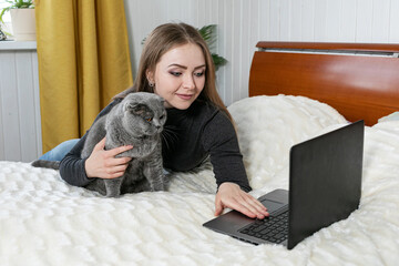 The girl lies on the bed with a laptop. Hugs a gray cat and looks into the laptop. Shopping for pets. Onlan shopping. Online training. Distance learning. We are sitting at home. Purebred cat