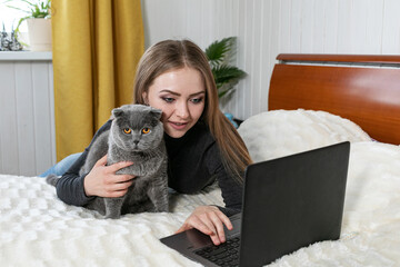 The girl lies on the bed with a laptop. Hugs a gray cat and looks into the laptop. Shopping for pets. Onlan shopping. Online training. Distance learning. We are sitting at home. Purebred cat