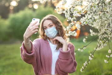A woman in a protective surgical mask, in pink clothes, with a phone in her hands, a masked model in a blooming garden.  allergy to flowering.  work during a pandemic.  Coronavirus disease, COVID-19