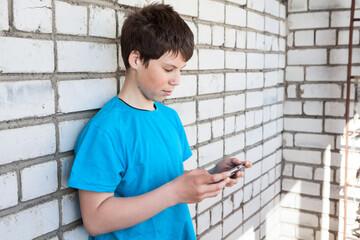 Obraz premium Attractive youngster guy wearing blue t-shirt holding smartphone in hands, browsing social media, leaning brick wall