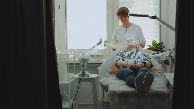 Beautician using vapozone facial steamer to make a professional facial skin treatment. Female client relaxing in beauty spa salon.