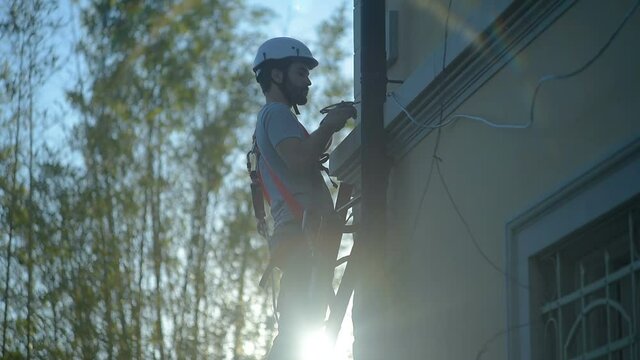 construction worker / laborer secures and tightens a cable standing on ladder on working building site, hard hat and safety equipment, sun flare on camera. Safety first at work concept