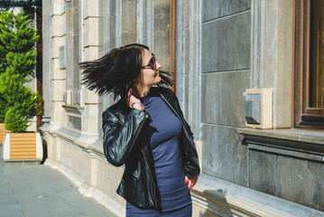 Brunette woman with a positive smile in a dress and leather black jacket shake hair in street and enjoys a sunny spring day. Girl travel in Batumi, Georgia. Old city at background.