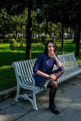 Nice, brunette woman in a dress sits on white bench in the street. Enjoys a sunny spring day. Girl traveling in Batumi, Georgia. Green park at background.