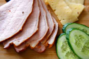 Slicing ham, cucumbers and cheese with ruddy thin breads lie on a wooden board. 