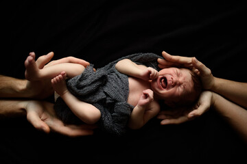 Little newborn boy on a black background. Top and bottom of the boy support the hands of parents. The baby is crying.