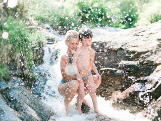A woman and a small child under a refreshing waterfall on a summer day. Blue swimsuit. Family day in the water. Water drops splashing the family