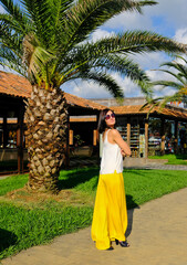 Romantic woman posing near big tree palm. Cheerful, smiling, lucky woman in white blouse, yellow trousers and sunglasses, outdoors. Sunny summer day.