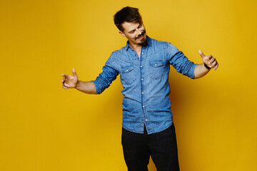 Fototapeta na wymiar Confident young handsome man in jeans shirt keeping arms wide and smiling while standing against a yellow background. Isolated with copy space.
