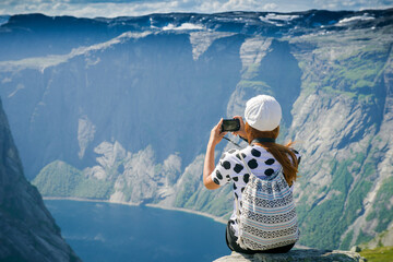 Girl tourist takes pictures. Girl sitting on the edge of a rocky cliff. Extreme tourism. Girl with a backpack. Mountains and fjords of Norway. tourist in a white cap on a cliff looks at the scenery