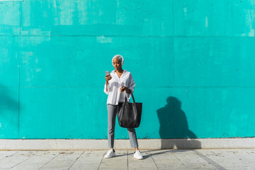 Businesswoman with headphones, standing in front of teal wall, using smartphone
