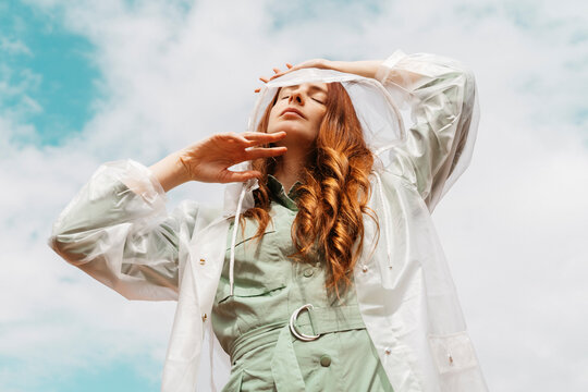 Redheaded Young Woman With Eyes Closed Wearing Transparent Rain Coat Standing Against Cloudy Sky