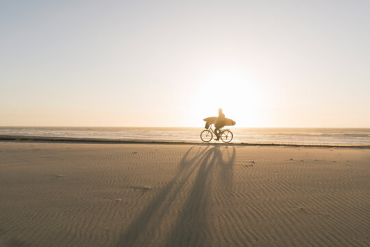 Surfer riding a bicycle during the sunset in the beach, Costa Nova, Portugal