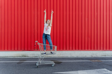 Girl in a shopping cart pointing fingers up in front of red wall