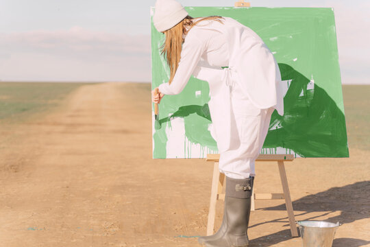 Young woman on dry field, painting canvas with green paint