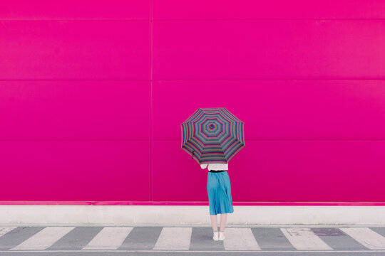 Rear view of young woman with umbrella standing in front of a pink wall