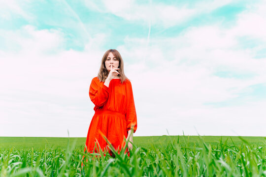 Portrait of woman wearing red dress standing in a field with finger on mouth