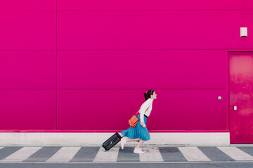 Young woman with smartphone running with trolley along a pink wall
