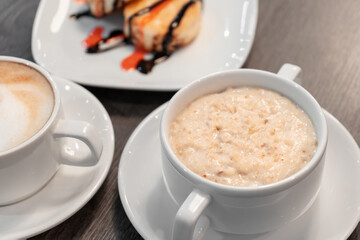 Oatmeal porridge on a wooden table near cheesecakes and cappuccino.