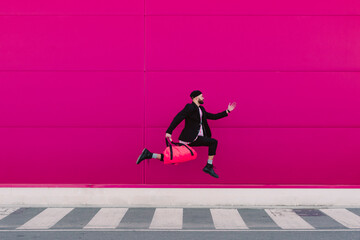 Young man jumping with travelling bag in front of a pink wall