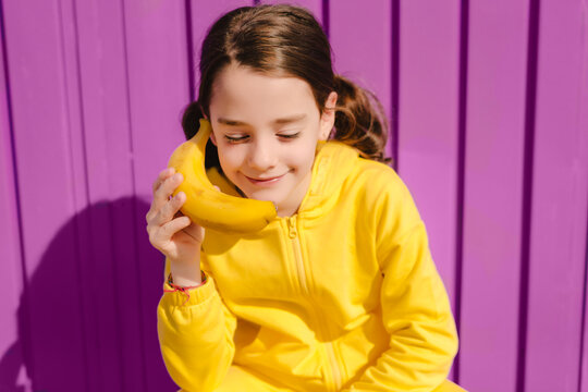 Portrait of smiling girl dressed in yellow holding banana in front of purple background