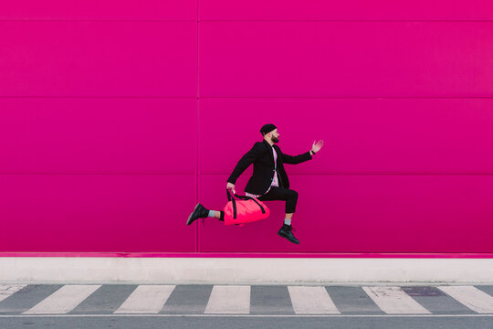 Young man jumping with travelling bag in front of a pink wall