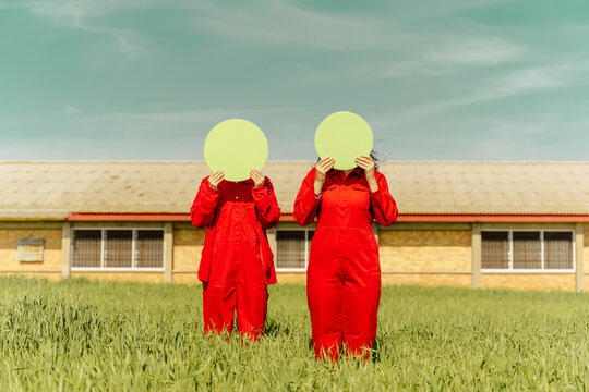 Young couple wearing red overalls  standing on a field hiding faces behind green circle