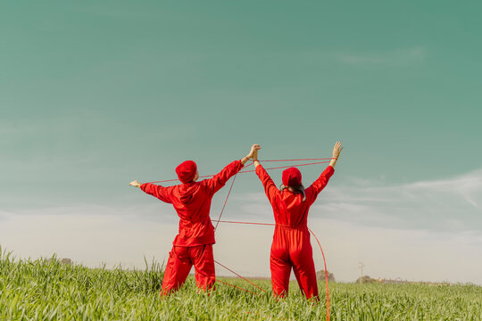 Back View Of Young Couple Wearing Red Overalls And Hats Performing On A Field With Red String