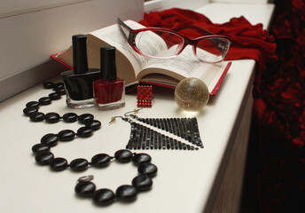 Naklejka premium Composition of red glasses, black beads made of natural agate, nail polishes, shiny disco earrings, a transparent shiny ball, an open dictionary, a matrix ring, on a white windowsill.