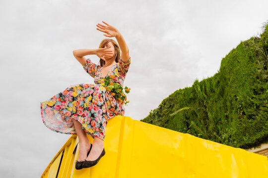 Woman in flower dress sitting on edge of yellow container with bucnh of flowers in her lap, moving arms