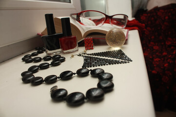 Composition of red glasses, black beads made of natural agate, nail polishes, shiny disco earrings, a transparent shiny ball, an open dictionary, a matrix ring, on a white windowsill.