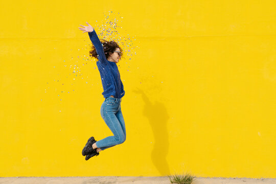 Young woman jumping in the air in front of yellow background throwing confetti
