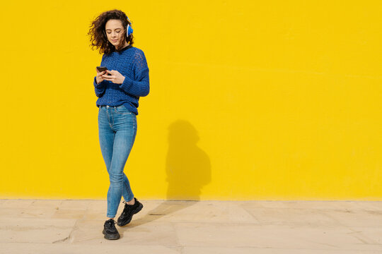 Portrait of young woman with headphones in front of yellow background looking at cell phone