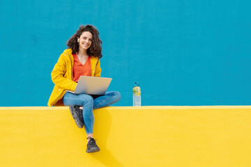 Portrait of smiling young woman with earphones and laptop sitting on yellow wall