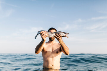 Young man standing in the sea holding caught fish in front of his face