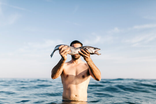 Young man standing in the sea holding caught fish in front of his face