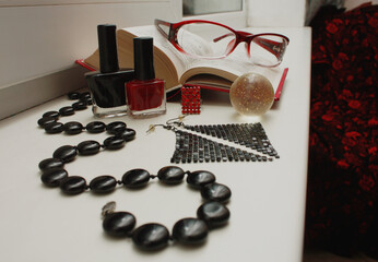 Composition of red glasses, black beads made of natural agate, nail polishes, shiny disco earrings, a transparent shiny ball, an open dictionary, a matrix ring, on a white windowsill.