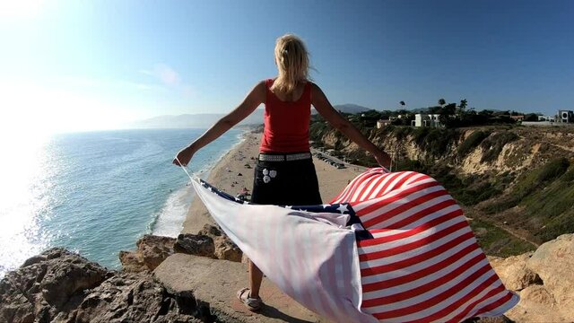 SLOW MOTION: Woman holding an American flag against the sun at Point Dume State Beach from Point Dume promontory on Malibu coast in CA, United States. Californian woman West Coast. Blue sunny sky.