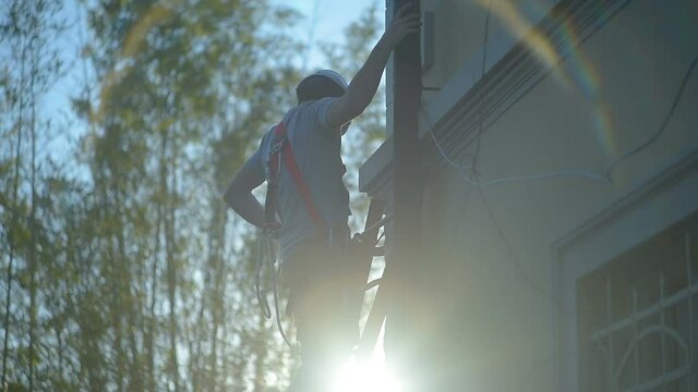 Young caucasian male builder worker/repairman climb down high ladder wearing protective white hardhat and safety equipment, golden hour sun ray flare, safety first at work education icon