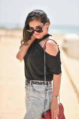 Sexy young woman on black t-shirt, fashion sunglasses and blue jeans with red fashion bag, stand on the beach. Girl doing sunbath on the beach sea.