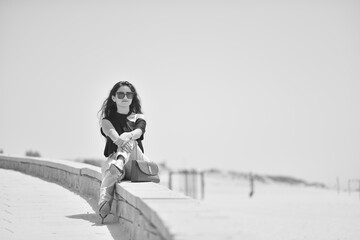 Sexy young woman on black t-shirt, fashion sunglasses and blue jeans sits on the beach, looking at the sea. Girl doing sunbath on the beach.